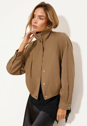 Frau mit hellbraunem Haar, die eine braune Jacke mit Knöpfen und einen schwarzen Rock mit Spitzenbesatz trägt, posiert mit Hand am Hals vor weißem Hintergrund.