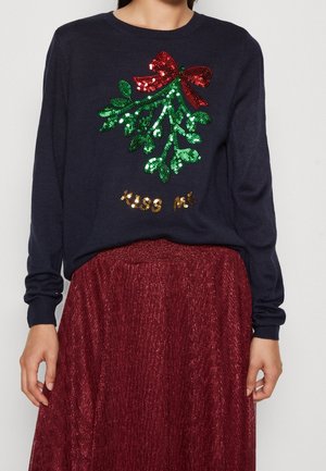 Pull bleu marine avec du gui vert en sequins et un nœud rouge au-dessus du texte doré en sequins « Kiss Me », assorti à une jupe rouge brillante.