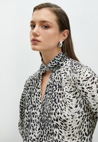 Svart och vit mönstrad blus med knuten halsringning, tillverkad av lättviktsmaterial. Modellen bär stora silverfärgade örhängen.