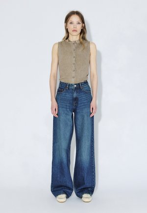 Beige sleeveless top with buttons and a rounded collar. Wide-leg blue jeans. Model wearing light-colored footwear. Neutral background.