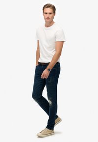 Camiseta de algodón blanca, jeans de mezclilla azul con detalles distress, zapatillas beige claro. Conjunto casual con un cinturón marrón y un reloj.