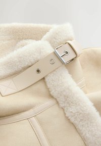 Beige schapenvacht kraag met een glad leren band, voorzien van een zilveren gesp en oogjes. Zachte textuur en warme uitstraling.