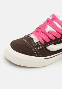 Bruna mockasiner i mocka med rosa skosnören, vita accentfärger och en texturerad vit gummisula. Sytt mönster ger extra detalj till designen.