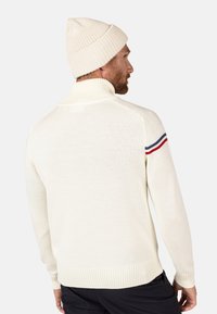 Maglione bianco lavorato a maglia con colletto alto, polsini e orlo a coste. Presenta una striscia rossa e blu sulla spalla sinistra. Abbinato a un berretto bianco.