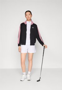 Zwarte en roze trainingsjack met rits, witte polo en witte shorts. Het model houdt een golfclub vast en draagt witte sneakers met zichtbare veters.
