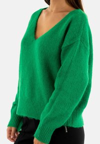 Maglione verde over-sized realizzato in materiale morbido e peloso. Presenta un profondo scollo a V, maniche lunghe e orlo a coste con una vestibilità rilassata.