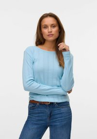 Lichtblauwe gebreide trui met ribbeltextuur en geometrische patronen, gecombineerd met donkerblauwe jeans en een bruine riem.