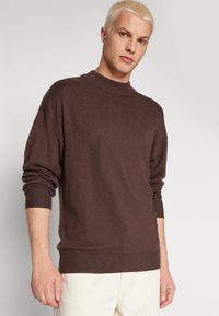 Jersey de punto marrón con cuello alto, puños de canalé y dobladillo. La tela parece suave y tiene una textura lisa.