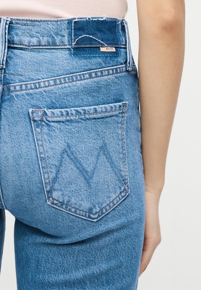 Οπίσθια όψη ατόμου που φοράει ψηλόμεσα μπλε τζιν με ραμμένο "M" στην πίσω τσέπη και μια μικρή ετικέτα "Mother" στη μέση.