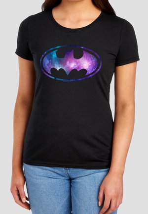 Frau trägt ein schwarzes T-Shirt mit buntem Galaxy-Batman-Logo und hellblauen Jeans und steht vor einfarbigem Hintergrund.