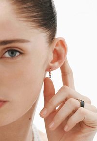 Pendiente de aro dorado con una pequeña piedra clara, usado en la oreja. Primer plano de una mano tocando la oreja; también son visibles anillos en los dedos.