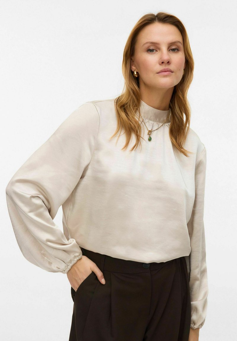 Licht beige blouse met lange, geplooide mouwen, een hoge halslijn en een gladde, licht glanzende textuur. Gecombineerd met donkere broek.