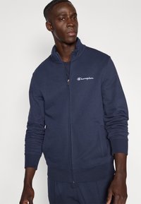 Μπλε σκούρο φλις ζακέτα με φερμουάρ, κατασκευασμένη από απαλό μείγμα βαμβακιού, με ψηλό γιακά, μακριά μανίκια και κεντημένο λογότυπο στο αριστερό στήθος.