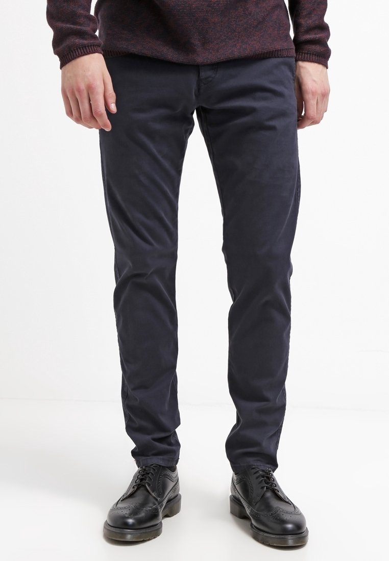 Homme portant un pantalon bleu marine foncé, des chaussures habillées en cuir noir et un pull bordeaux, debout sur un fond blanc.