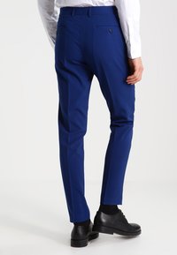 Blå skräddarsydda byxor i mjukt tyg, med bakfickor med knappar, slim fit ben, och i kombination med svarta skor.