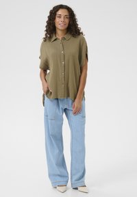 Olive green, short-sleeve shirt, textured fabric, button front. Paired with light blue wide-leg jeans and cream-colored shoes.
