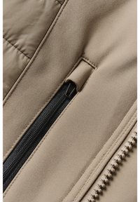 Gros plan sur une veste beige avec une poche noire zippée et des coutures apparentes, montrant la texture du tissu et une fermeture éclair beige sur le bord.