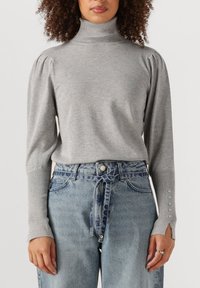 Grauer gestrickter Rollkragenpullover mit Puffärmeln und Knopfdetails, kombiniert mit hoch-taillierten hellblauen Jeans. Texturiertes Gewebe und entspannter Schnitt.