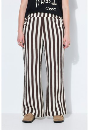 Person iført sorte og hvide lodrette stribede vidde bukser, sort skjorte, ringe og brune slip-on sko står mod en hvid baggrund.