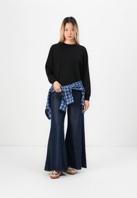 Zwarte longsleeve sweatshirt, blauwe geruite flannel om de taille geknoopt, wijde donkerdenim jeans en beige schoeisel. Eenvoudig en casual ontwerp.