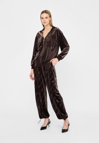 Chándal de terciopelo marrón con sudadera con cremallera que cuenta con dos bolsillos en el pecho, acompañado de pantalones ajustados a juego, decorados con un patrón de logotipos.
