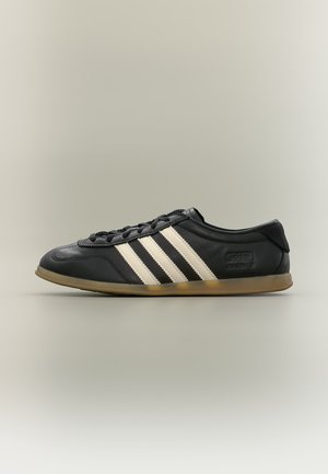 Μαύρο αθλητικό παπούτσι Adidas Gazelle με λευκές ρίγες, μαύρα κορδόνια και ανοιχτό καφέ σόλα, εμφανιζόμενο σε πλάγια όψη σε ουδέτερο φόντο.
