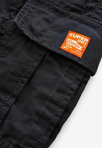 Čierne látkové šortky s viditeľným šitím, s výraznou oranžovou nálepkou "Superdry" na vrecku. Textúrovaný materiál s utilitárnym dizajnom.