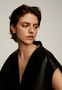 Prenda de cuero negro con un profundo escote en V y mangas cortas, llevada sobre un delicado collar de oro visible y pendientes plateados redondos.