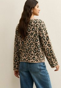 Beige trui met luipaardprint en donkergroene vlekken, met een comfortabele pasvorm en verlaagde schouders, gecombineerd met een blauwe jeans.