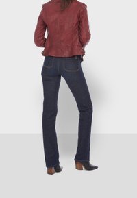 Rote Lederjacke mit asymmetrischem Saum, kombiniert mit dunkelblauen Jeans. Das Modell trägt schwarze Absatzstiefel, die eine figurbetonte Silhouette zeigen.