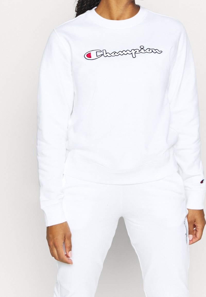 Sweatshirt blanc en polaire, avec un col rond et des poignets côtelés, arborant un logo "Champion" brodé en noir et rouge bien visible.