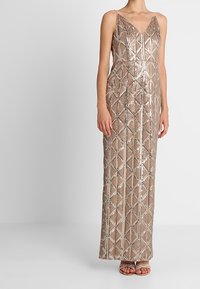 Abito senza maniche, lungo fino al pavimento, di colore beige, con un motivo geometrico di paillettes lucide, scollatura a V, indossato con sandali con tacco alto bianchi e con cinturini.