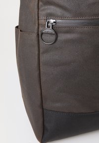 Brown fabric bag corner with black bottom panel and Barbour-branded metal zipper pull on front pocket.
