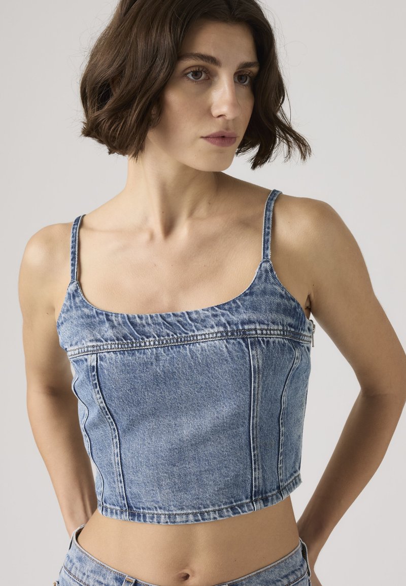 Donna con capelli castani corti che indossa un top corto di jeans blu senza maniche, che guarda di lato contro uno sfondo semplice.