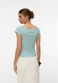 Vrouw met haar in een knot, gekleed in een lichtblauw topje met korte mouwen en een witte rok, staand met haar rug naar de camera tegen een effen achtergrond.