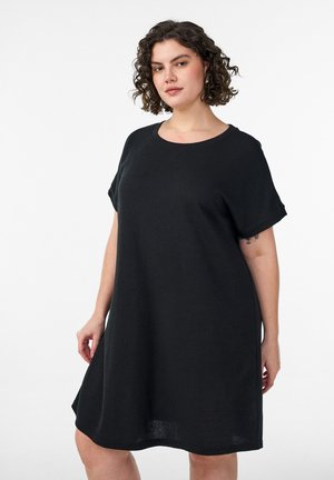 Žena s kovrčavom kosom u širokoj crnoj haljini s kratkim rukavima, stoji na jednobojnoj bijeloj pozadini.