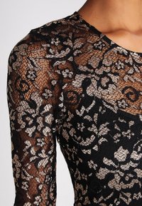 Top en dentelle noire avec des motifs floraux intriqués et une texture transparente. Manches longues avec un col arrondi, mettant en valeur des éléments de design superposés.