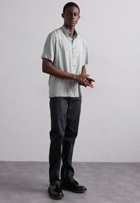 Chemise à manches courtes, à carreaux verts et blancs, pantalon noir et chaussures noires, se tenant contre un fond uni. Mains jointes.