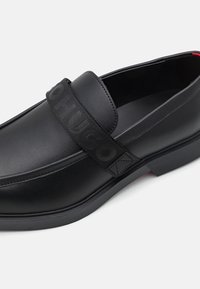Fekete bőr, slip-on cipő széles, "HUGO" felirattal ellátott elasztikus szalaggal. Kerek orrú és texturált gumitalpú, amely biztosítja a tapadást.