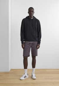 Schwarzer Kapuzenpullover mit Kordeln, lila Shorts und weißen Sneakern mit schwarzen Streifen. Das Model steht auf einem Holzboden in einer minimalistischen Umgebung.