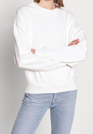 Witte oversized sweatshirt met een ronde hals en verlaagde schoudernaden; zachte textuur, geribde manchetten en zoom, gecombineerd met lichtblauwe jeans.