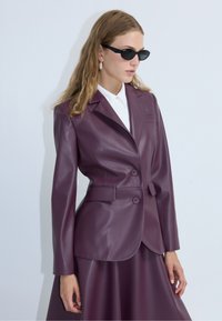 Blazer en simili cuir violet avec revers crantés, deux boutons et poches latérales. Porté sur une chemise blanche à col, texture lisse, design ajusté.