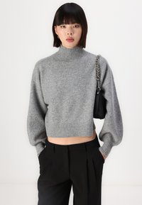 Grå cropped genser med høy ribbestrikket hals og ballongsleeves, laget av mykt, teksturert materiale. Kombinert med svarte plissert bukser.