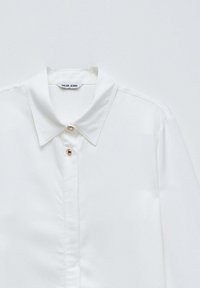 Camisa blanca de cuello con dos botones dorados visibles y una etiqueta "SALSA JEANS" sobre un fondo claro y sencillo.