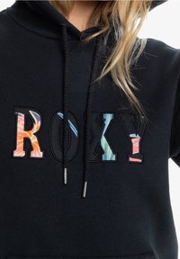 Sweat à capuche noir arborant "ROXY" en lettres épaisses ; les lettres affichent un motif floral en orange, rose et bleu avec des coutures texturées.