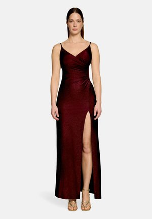 Frau steht und trägt ein ärmelloses, bodenlanges bordeauxrotes Kleid mit hohem Schlitz am linken Bein und goldenen offenen Zehenabsätzen.