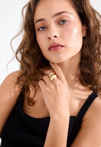 Gold-colored ring with a smooth, curved design featuring layered bands. Model has wavy, shoulder-length hair and wears a black top.
