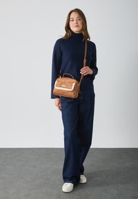Sac à main en suède beige avec une touche de fourrure crème. Le sac possède une poignet supérieur et une bandoulière ajustable, avec une forme rectangulaire et du matériel doré.