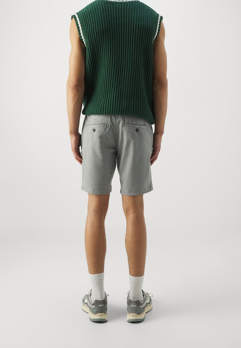 Zelený pletený bezrukávový top, šedé šortky a šedé tenisky s akcenty, viděno z pohledu zezadu. Top má hrubou texturu a bílý lem.