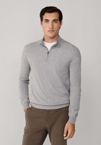 Joven vestido con un suéter gris claro de media cremallera sobre una camisa blanca y pantalones marrones, de pie con una mano en el bolsillo frente a un fondo liso.
